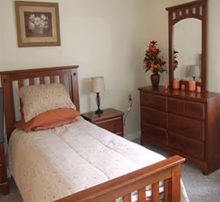 A bedroom with a single wooden bed featuring a floral-patterned bedspread and pillows. Next to the bed is a wooden nightstand with a lamp. Across from the bed is a wooden dresser with a large mirror, decorated with a vase of flowers and several candles. A framed floral painting hangs on the wall above the bed.