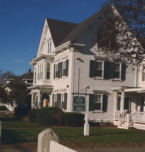 A large white multi-story house with green shutters and a steep roof, surrounded by bushes and a white picket fence. A sign in front reads 'Seth Mann 2nd Home'. The sky is clear and blue.