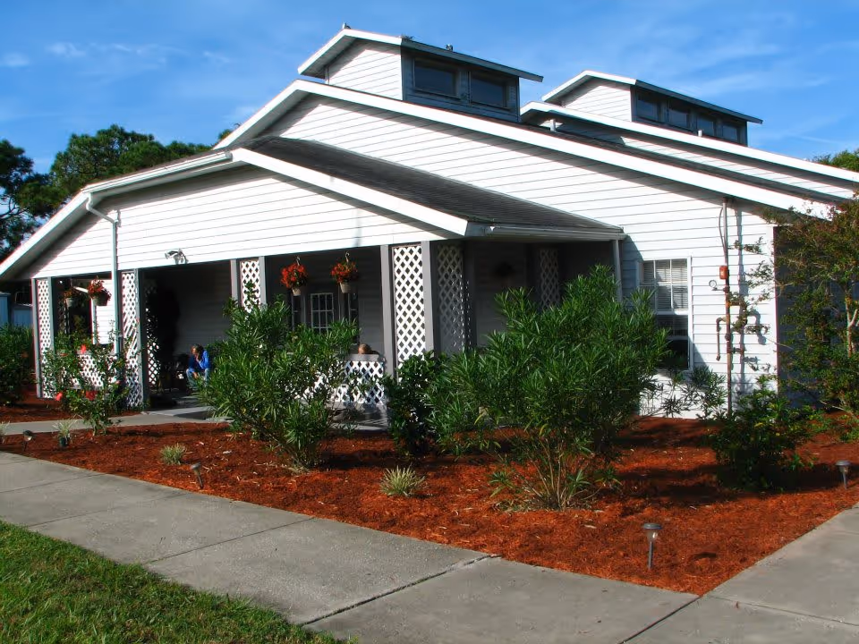 Exterior view of a white single-story building with a covered porch, lattice columns, hanging flower pots, and landscaped garden beds with shrubs and mulch. A concrete sidewalk runs along the front of the building under a clear blue sky.