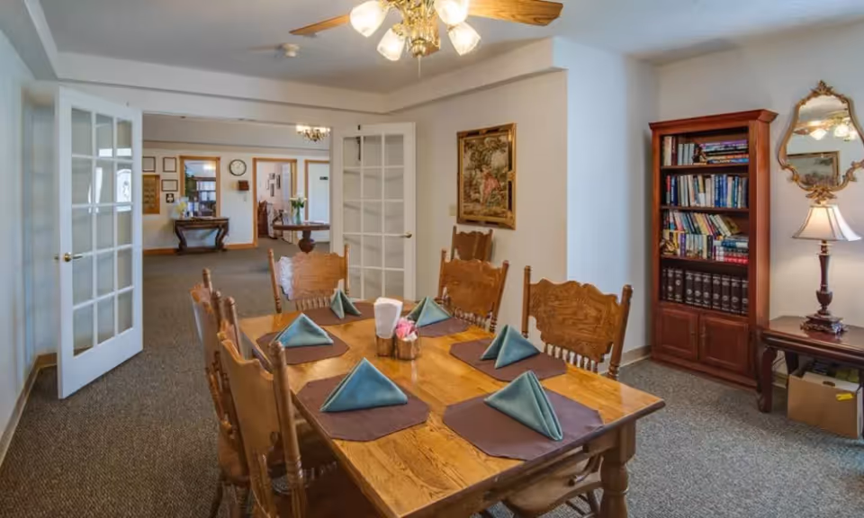 A dining area with a wooden table set for six people with folded blue napkins on brown placemats. The room has wooden chairs, a ceiling fan with lights, a bookshelf filled with books, a lamp on a side table, a decorative mirror, and a framed painting on the wall. French doors open into another room with additional furniture and decor.