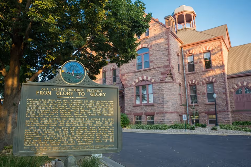 Historic stone building with arched windows and a small tower, partially shaded by a large tree. In front of the building is a green historical marker sign titled 'All Saints Historic District From Glory to Glory' with detailed text about the history of the site.
