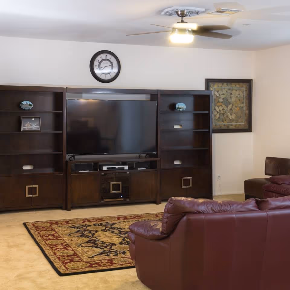 A living room with a large flat-screen TV mounted on a dark wooden entertainment center with shelves and cabinets. There is a decorative rug on the beige carpeted floor and two maroon leather sofas facing the TV. A ceiling fan with a light is on the ceiling, and a framed artwork hangs on the wall to the right.