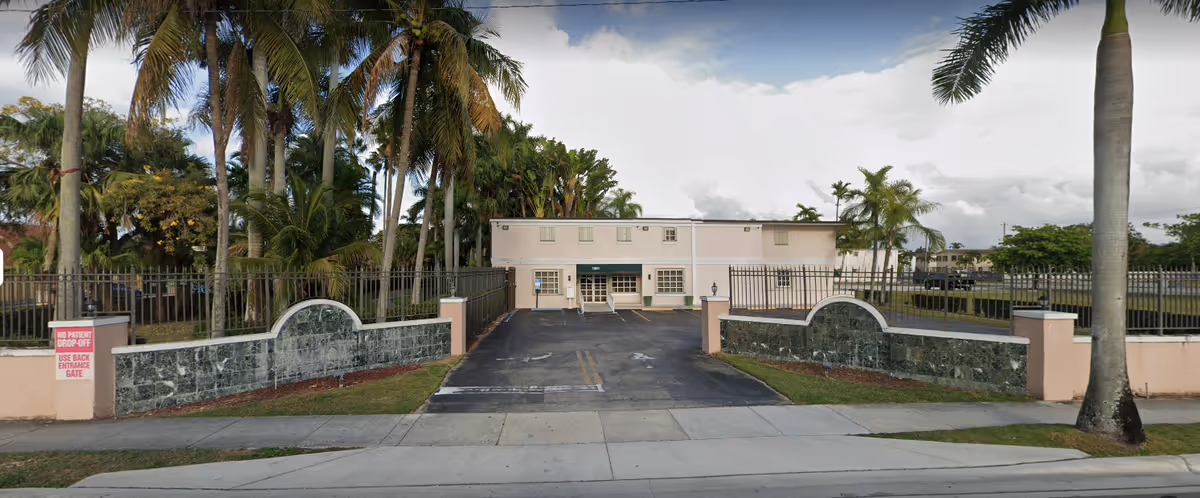 Front exterior of a gated, two-story institutional building with a driveway, iron fencing and palm trees.