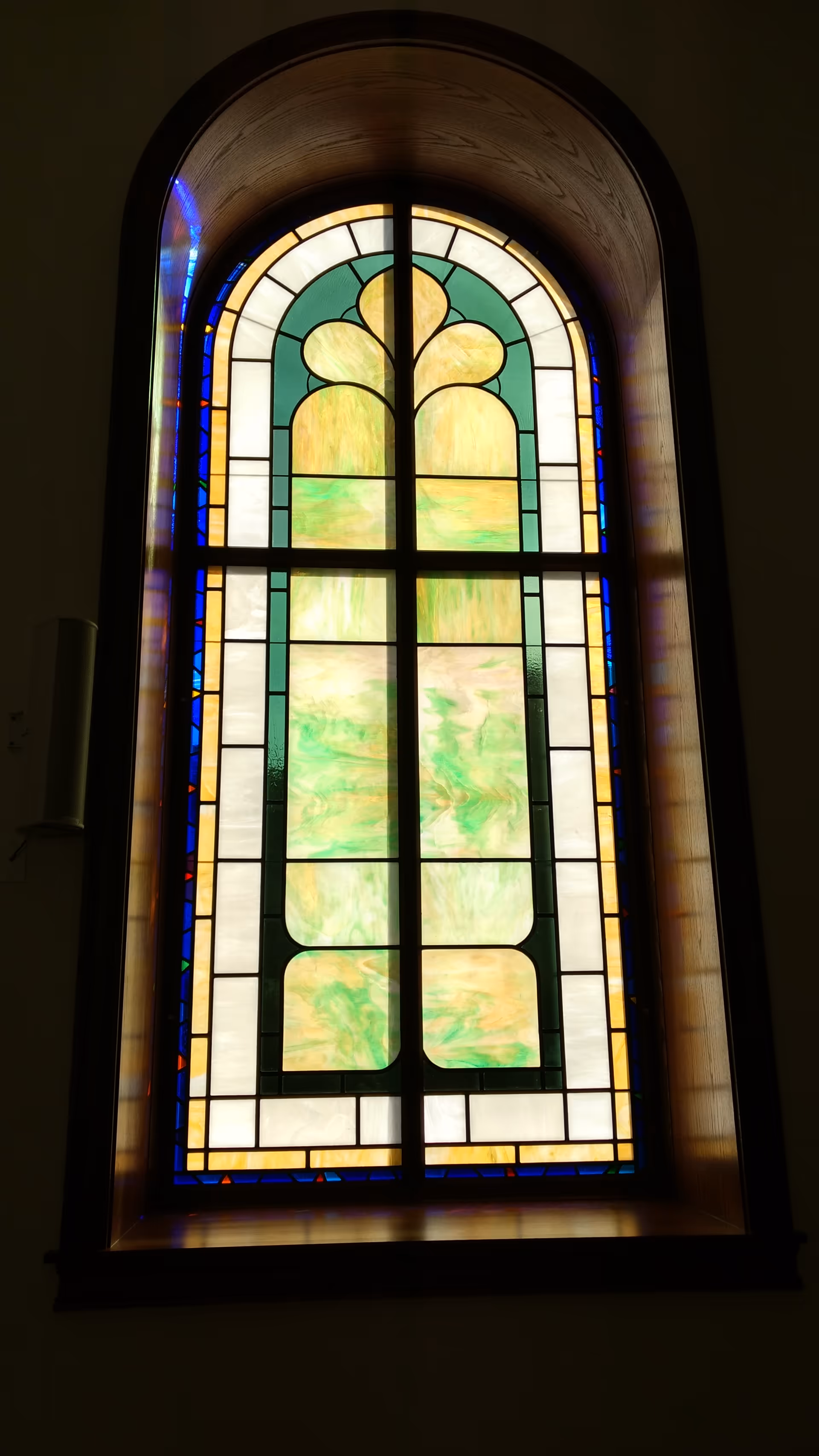 Tall arched stained glass window with geometric panes in green, gold, and blue indoors.