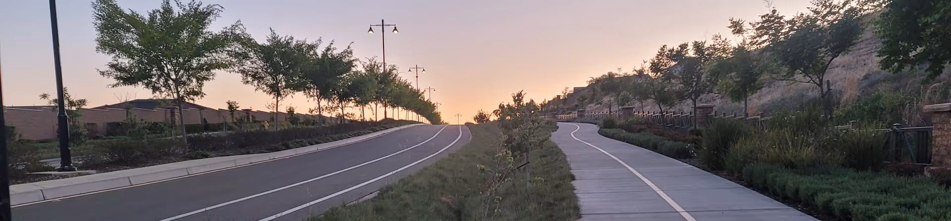 Sunset-lit avenue with a winding concrete path, trees, and streetlamps along landscaped slopes.