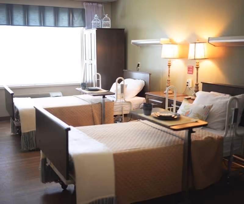 Two hospital-style beds in a softly lit senior living bedroom with bedside tables, lamps, and overbed trays.