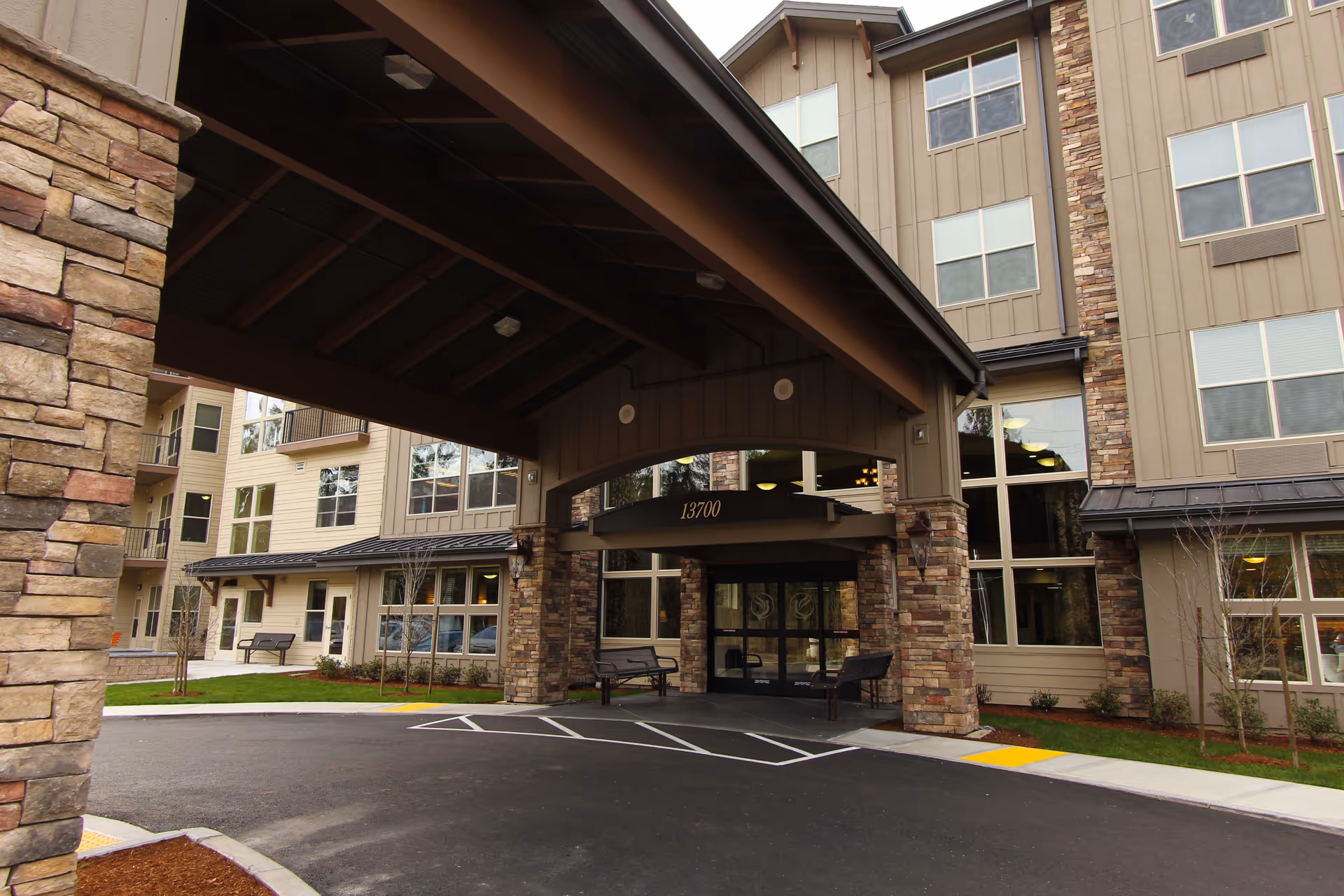 Entrance of a senior living facility with a covered drop-off area supported by stone pillars, large windows, and a building number 13700 above the entrance doors. The building exterior features beige siding and stone accents with benches near the entrance.