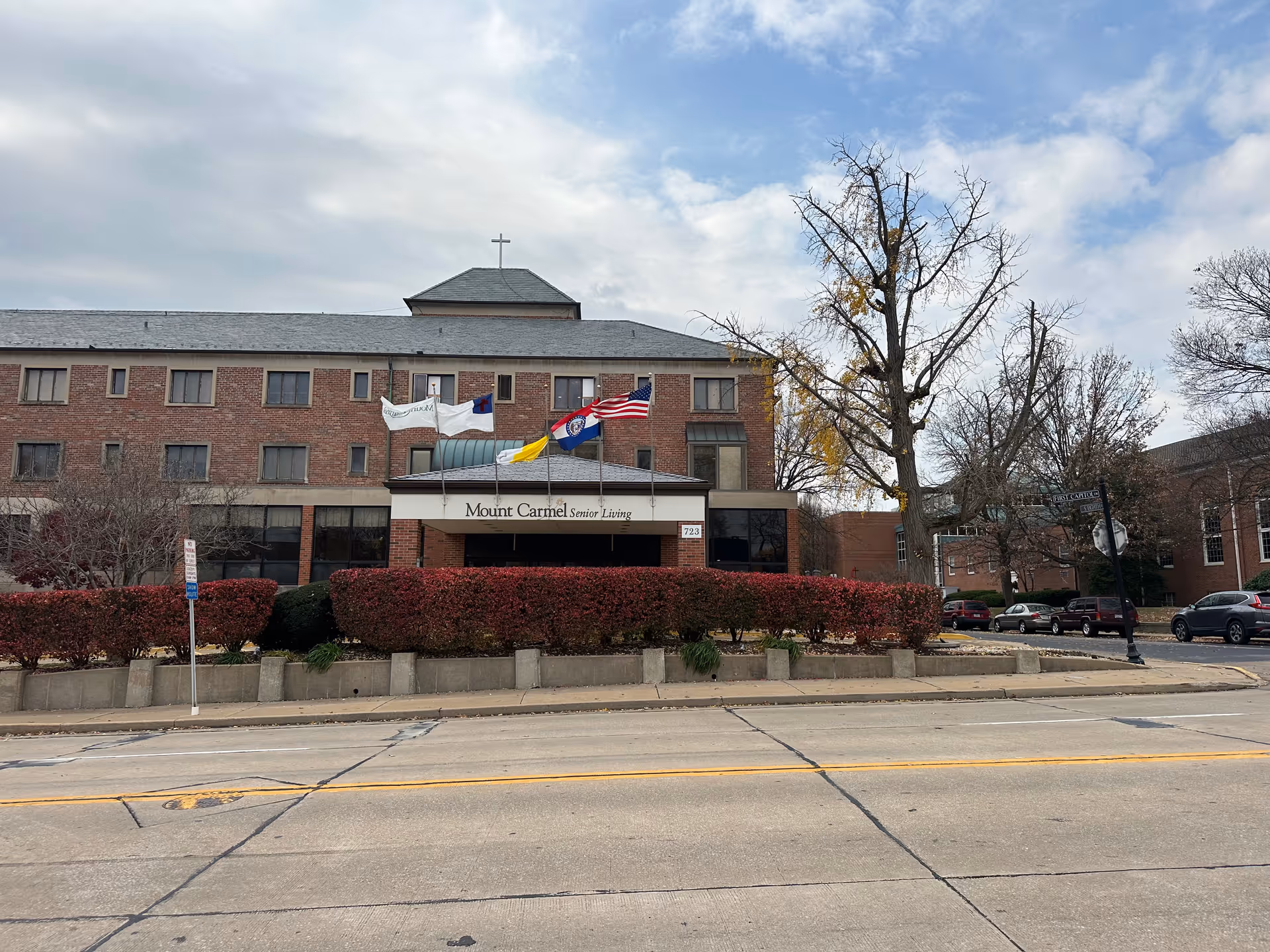 Brick facade of Mount Carmel Senior Living with an entrance canopy, flags, and landscaped hedges along the street.