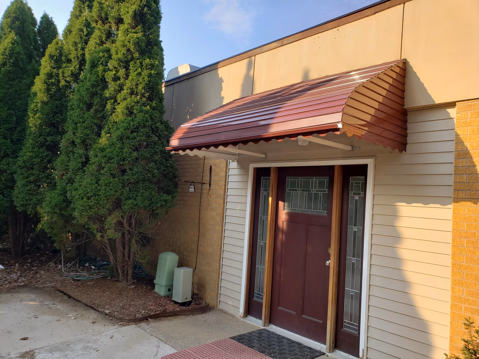 Exterior entrance with maroon door, red awning, beige siding, and tall evergreen trees.