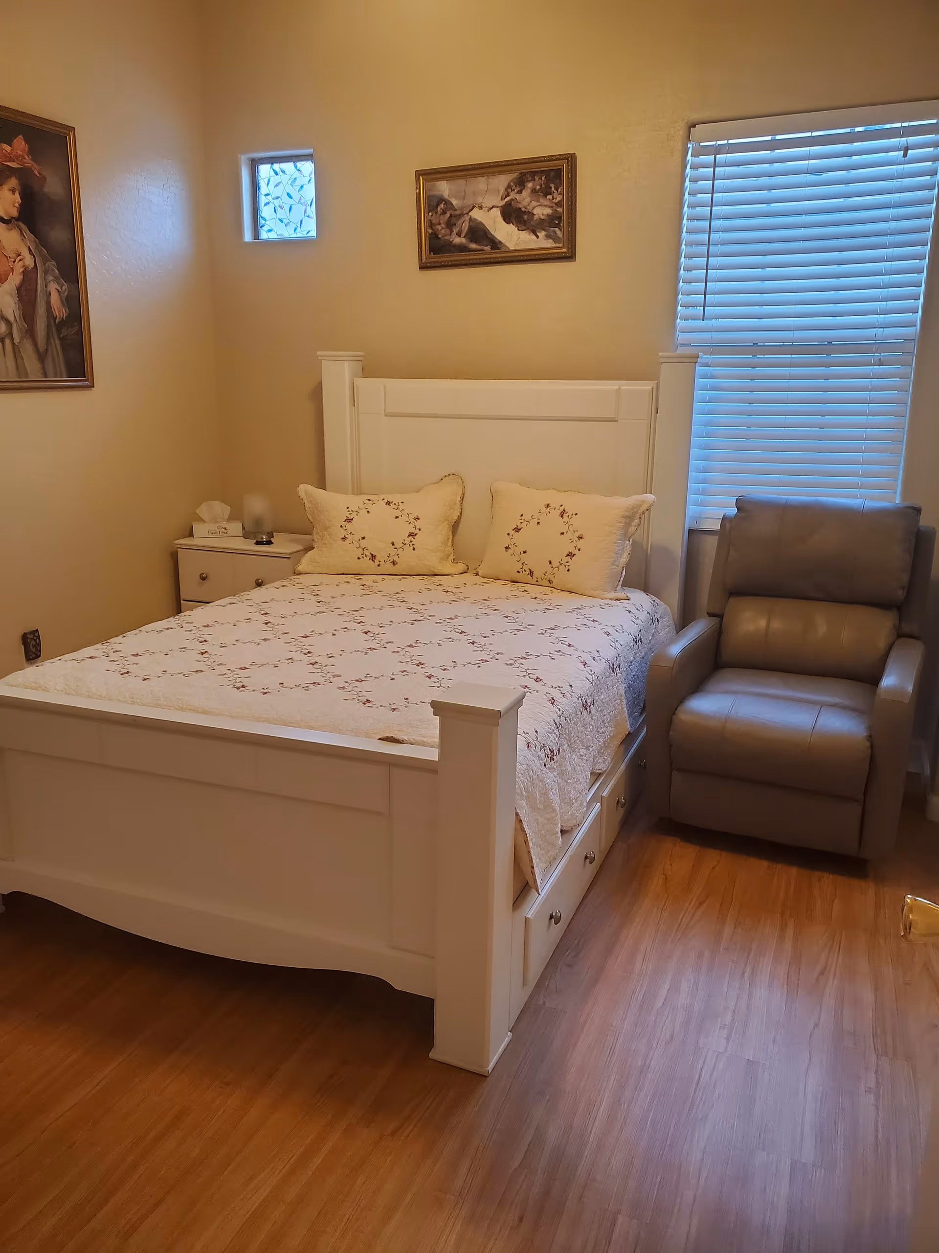 A cozy bedroom with a white wooden bed frame and a floral quilt. Two matching pillows are placed on the bed. To the right of the bed is a gray recliner chair next to a window with closed blinds. On the left side of the bed is a small white nightstand with a tissue box and a small lamp. The walls are beige and decorated with two framed paintings. The floor is wooden.