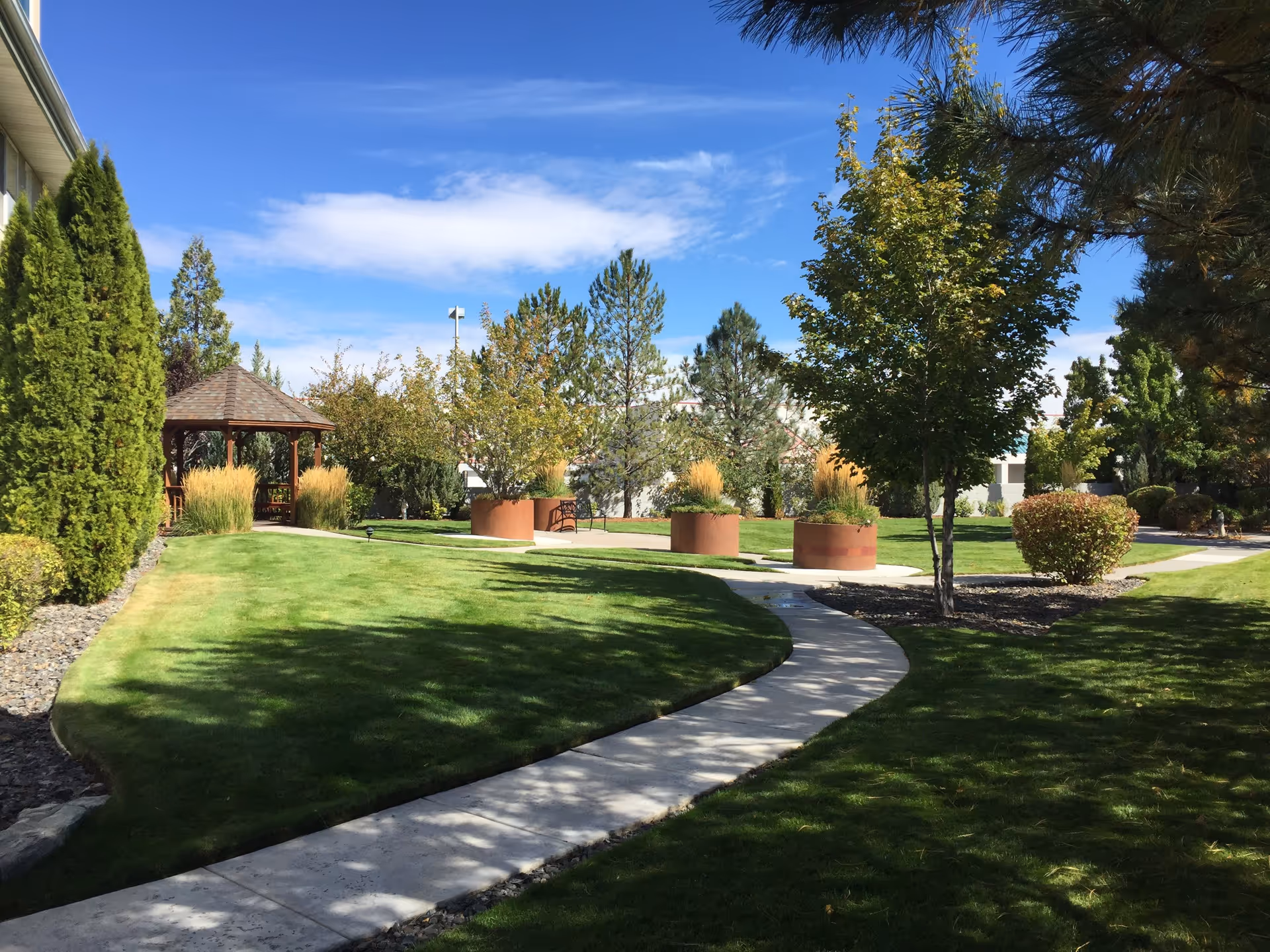 A well-maintained outdoor garden area at Sierra Place Retirement Community featuring a curved concrete walkway, green grass, various trees and shrubs, large round planters with ornamental grasses, and a wooden gazebo under a blue sky with some clouds.