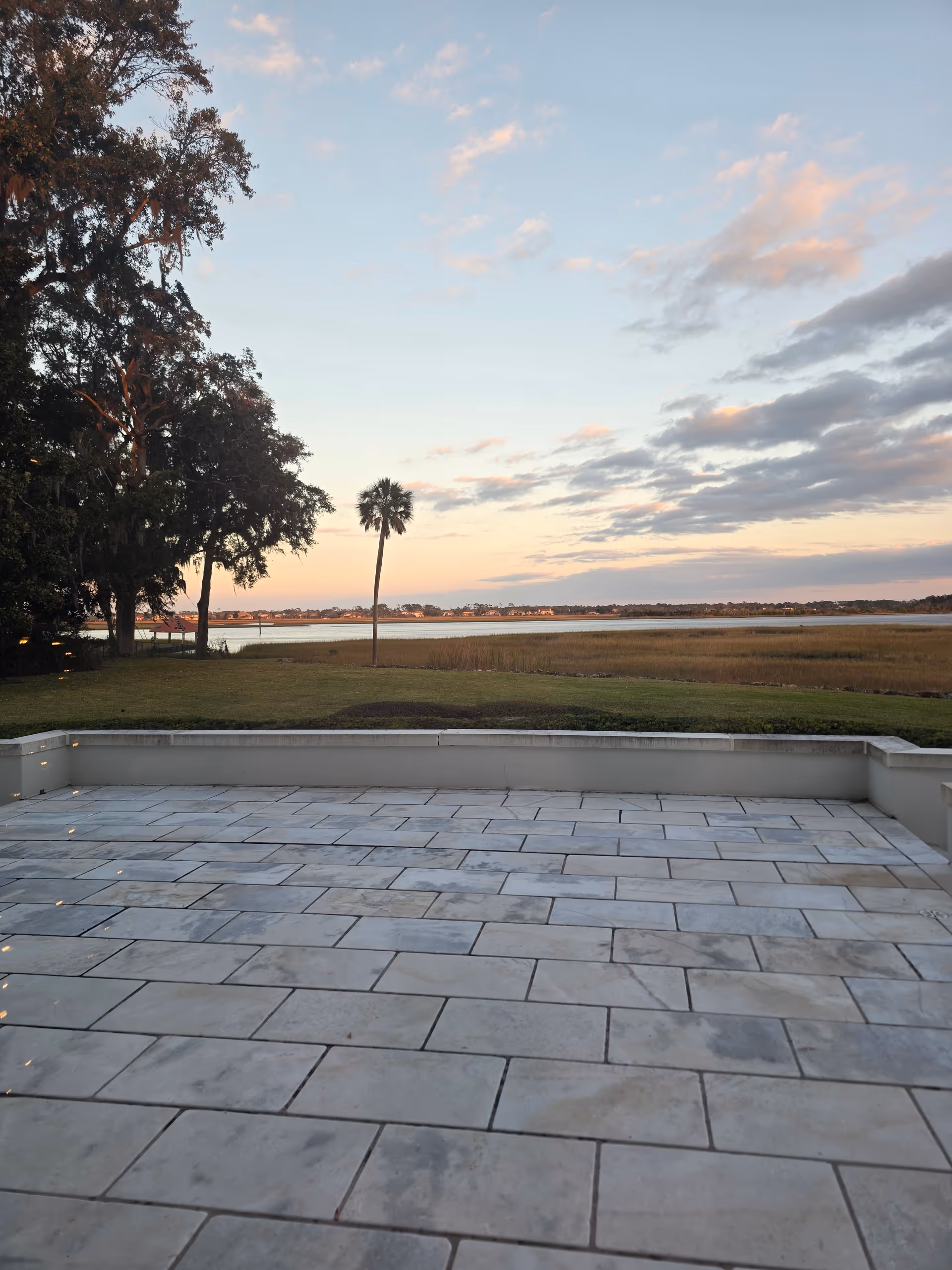 Sunset over calm water visible beyond a tiled outdoor terrace with trees.