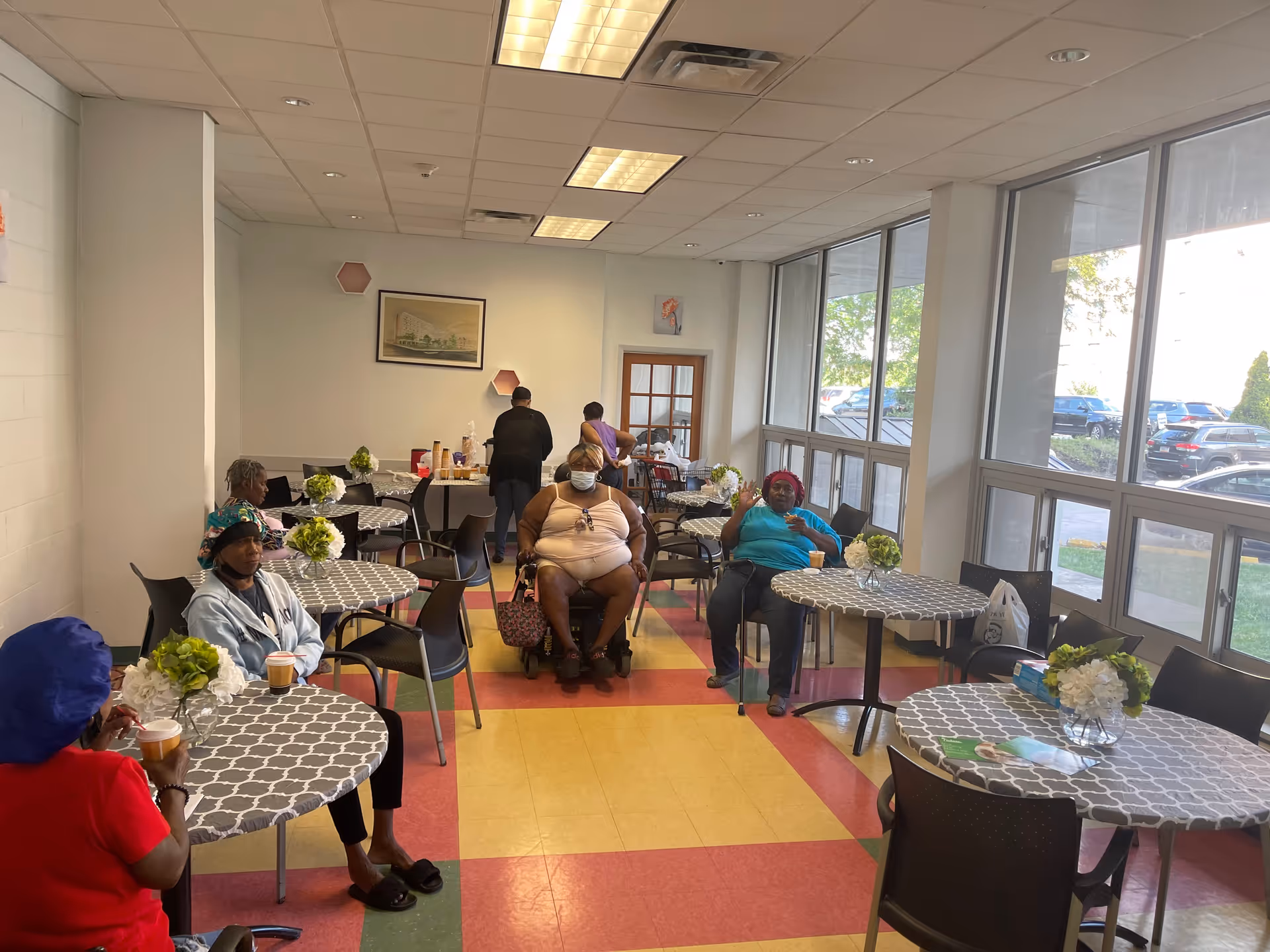 Residents sit at round tables with checkered tablecloths in a bright communal dining area.