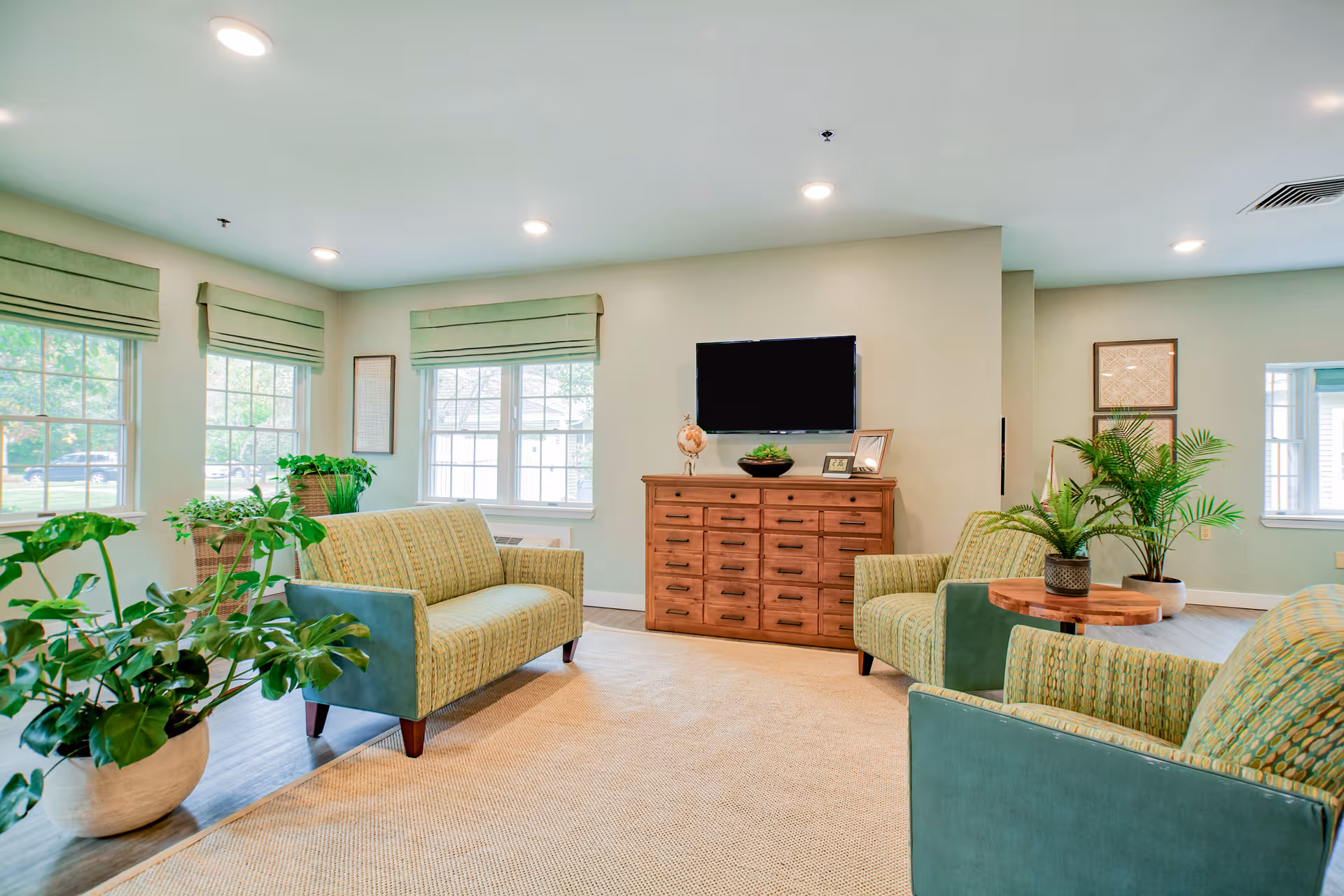 Bright lounge area with green upholstered seating, plants, and a wooden dresser under a mounted TV.