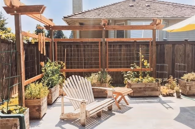 Sunny backyard patio with a wooden pergola, planter boxes, a white Adirondack chair and small side table.