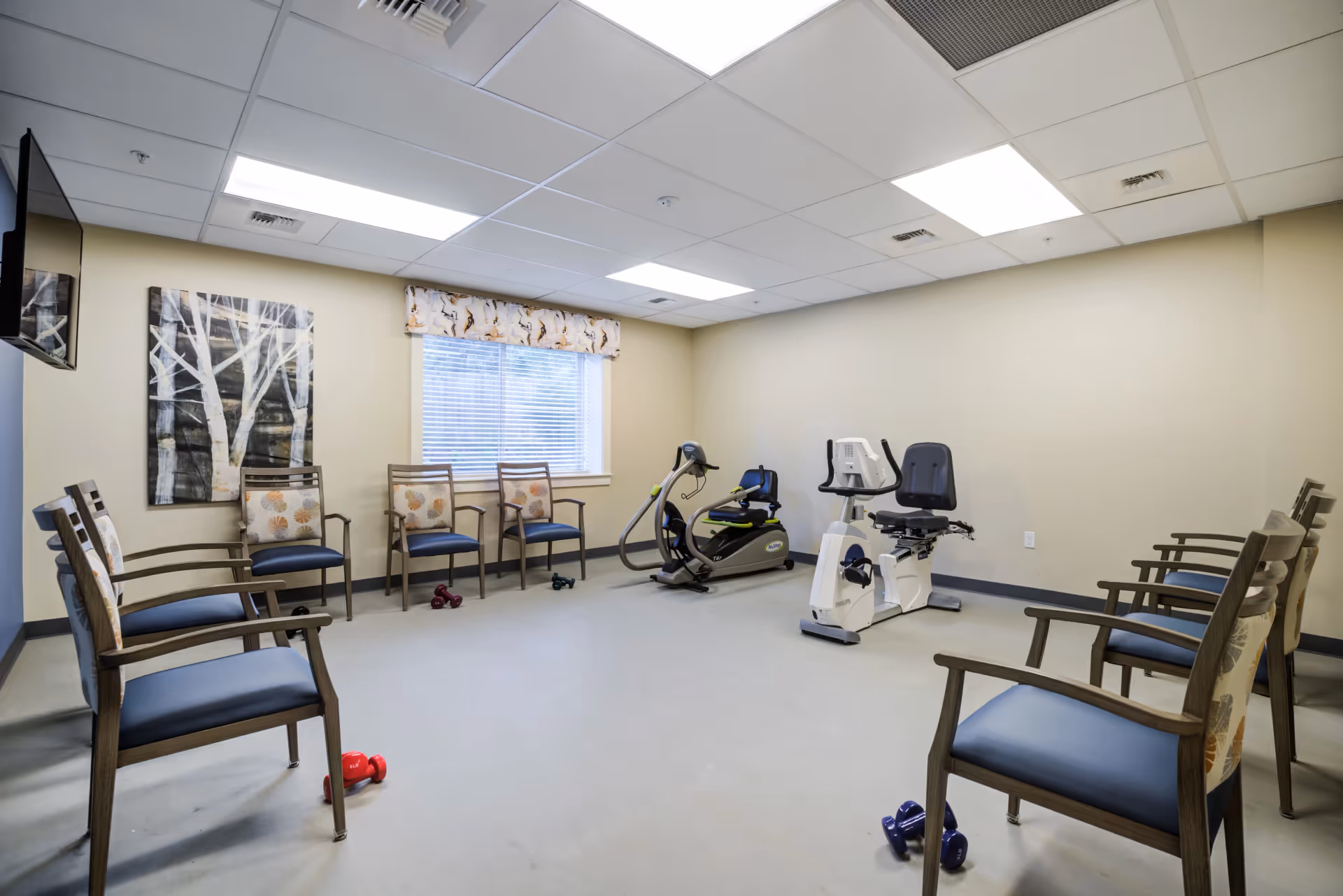 Well-lit senior exercise room with chairs around the walls, a window, two recumbent exercise bikes, and small dumbbells on the floor.