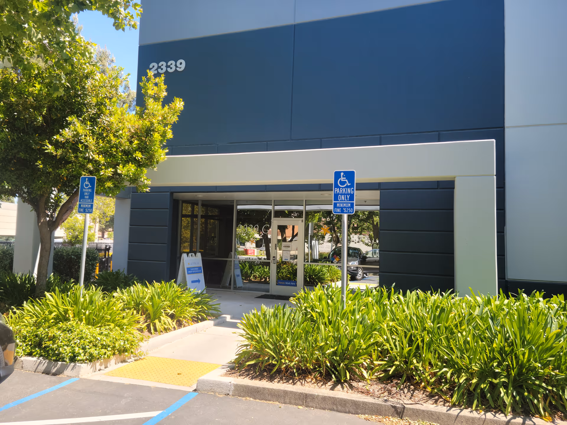 Exterior view of a modern building entrance with glass doors, plants, and handicap signage.