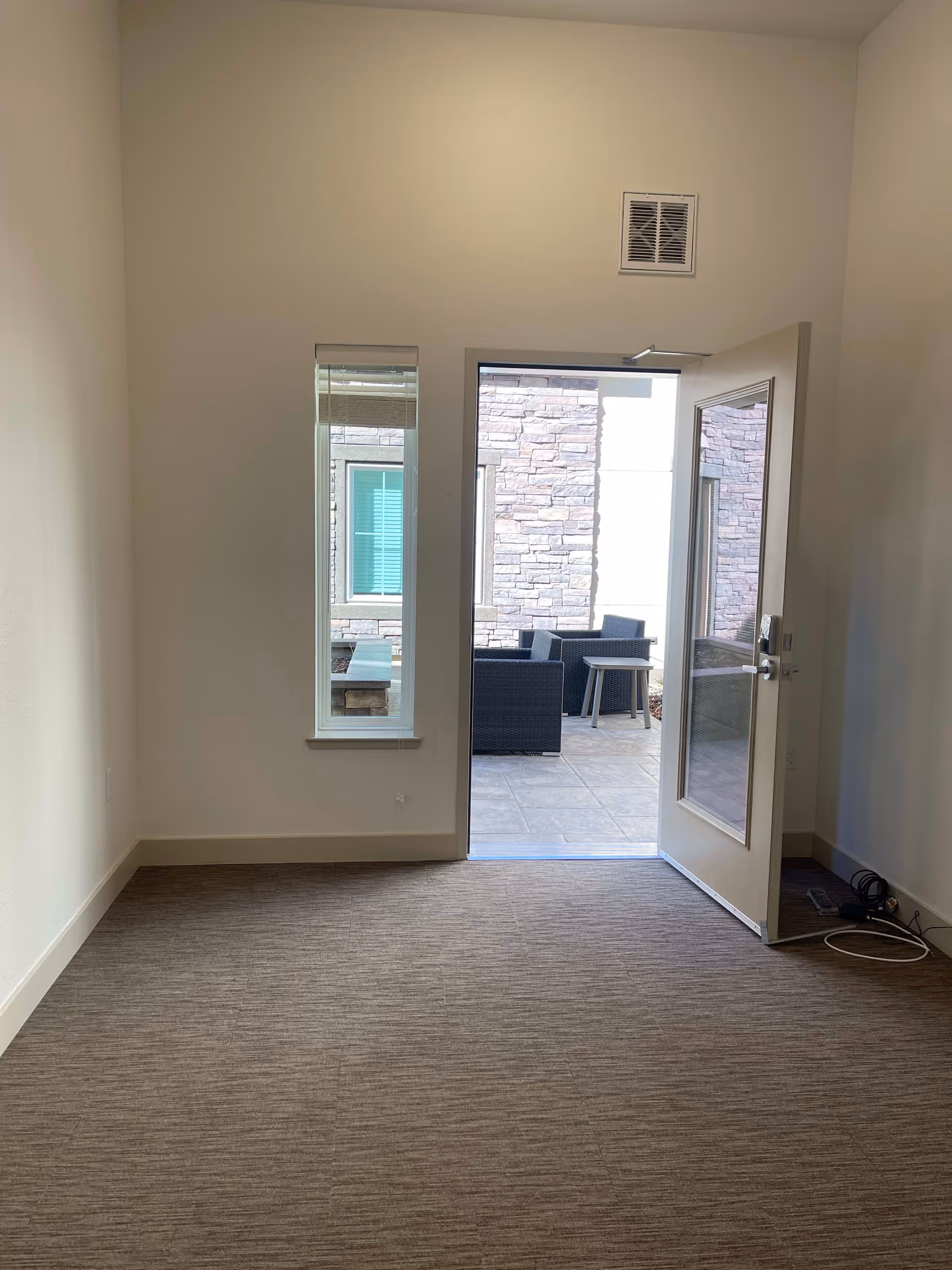 Empty room with beige walls and carpeted floor, featuring a narrow vertical window and an open door leading to an outdoor patio area with stone walls and black wicker furniture including chairs and a small table.