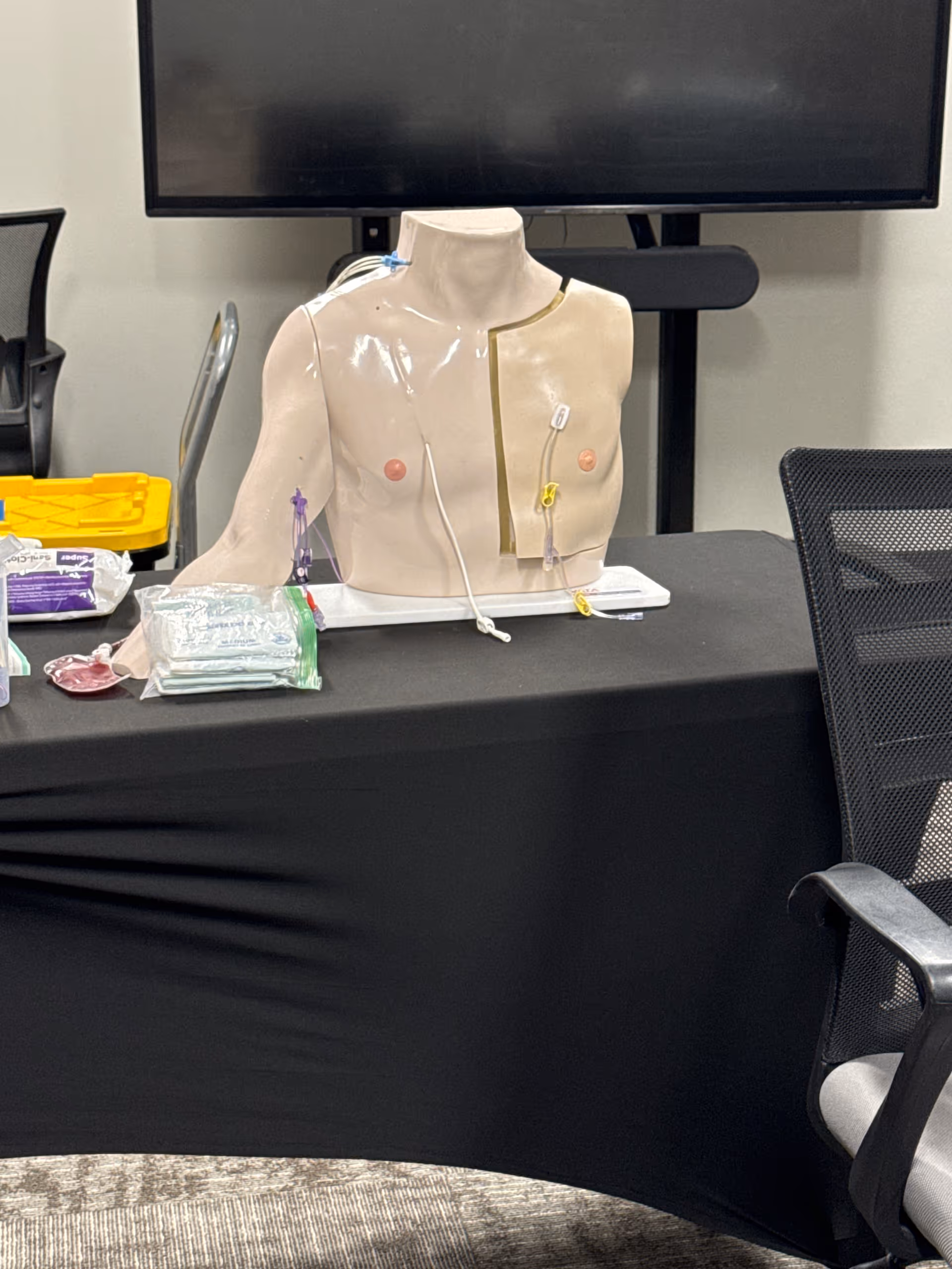 Medical training mannequin torso with wires and supplies on a table indoors.