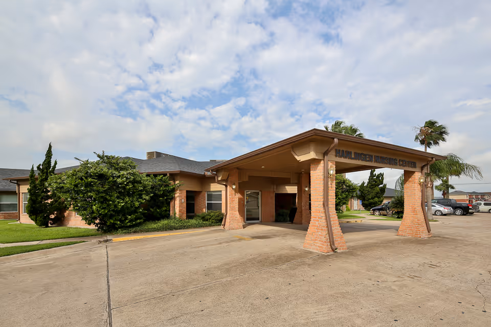 Exterior view of Harlingen Nursing and Rehabilitation Center showing the entrance with a covered driveway supported by brick pillars, surrounded by greenery and parked cars under a partly cloudy sky.