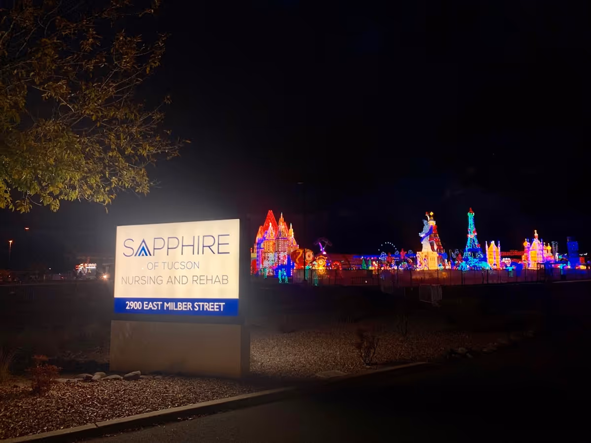 Illuminated exterior sign for Sapphire of Tucson Nursing and Rehab at night with colorful holiday light displays in the background.