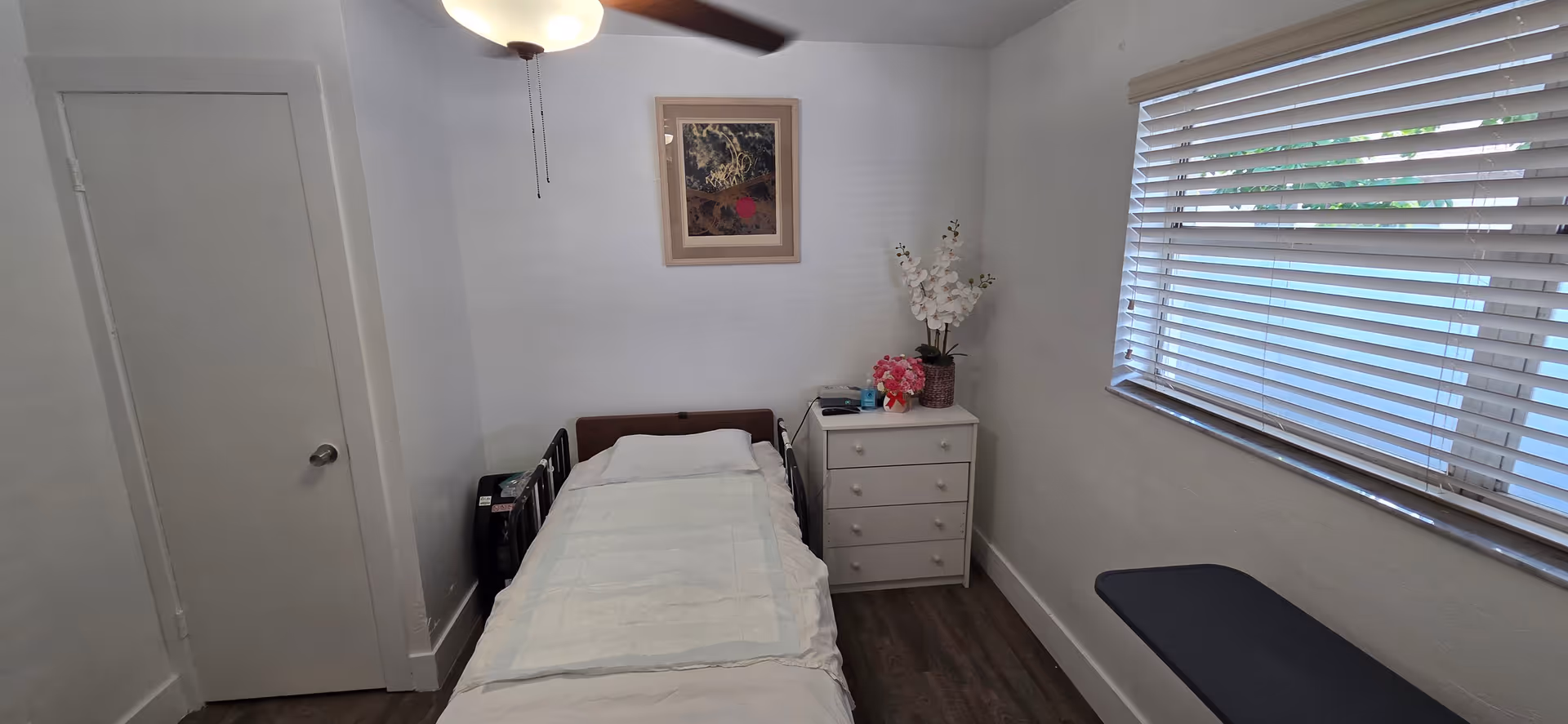 Small, neatly made bedroom with single bed, dresser, and large window blinds.