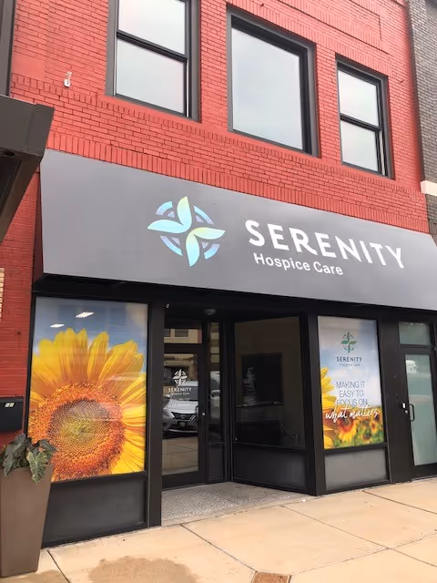Red brick building storefront with Serenity Hospice Care sign and sunflower posters at entrance