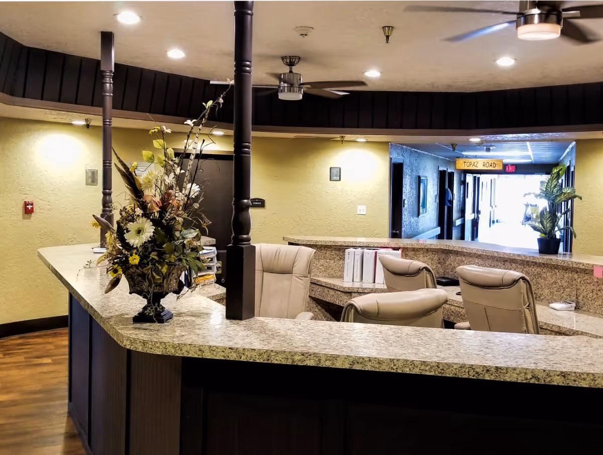 Reception desk area inside a nursing home with a granite countertop, beige office chairs, a large floral arrangement, and a hallway in the background with a sign that reads 'Topaz Road'. The walls are painted light yellow and the floor is wooden.
