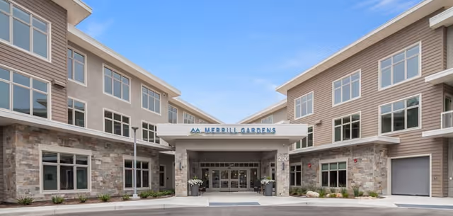 Front entrance of Merrill Gardens at Monterey showing a three-story building with stone and siding facade, large windows, and a covered drop-off canopy.