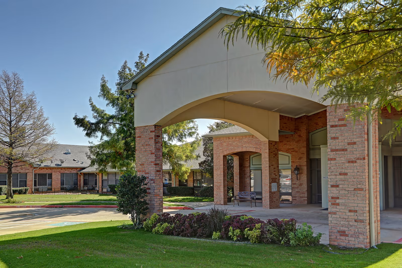 Exterior view of a senior living facility with a covered entrance supported by brick columns. There is a well-maintained lawn with shrubs and trees around the building, and a bench is visible under the covered area.