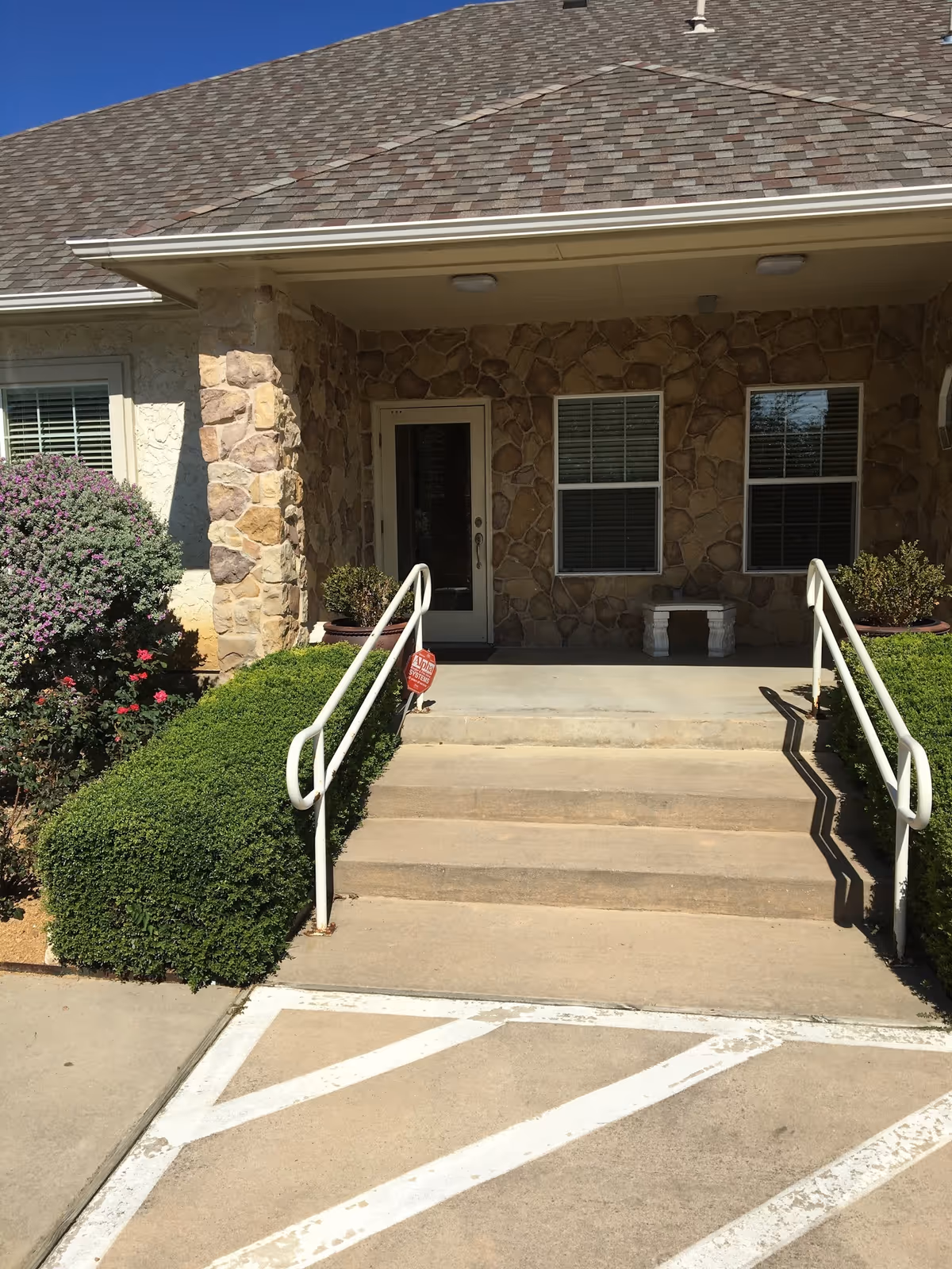 Front entrance of a stone-faced senior living facility with steps, railings, and shrubs.