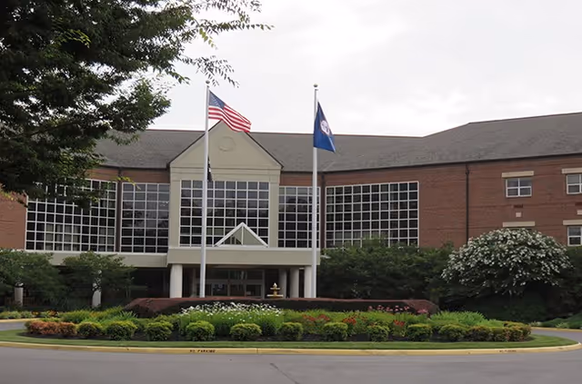 Front entrance of a brick care facility with a large glass facade, two flagpoles, and a landscaped circular driveway.
