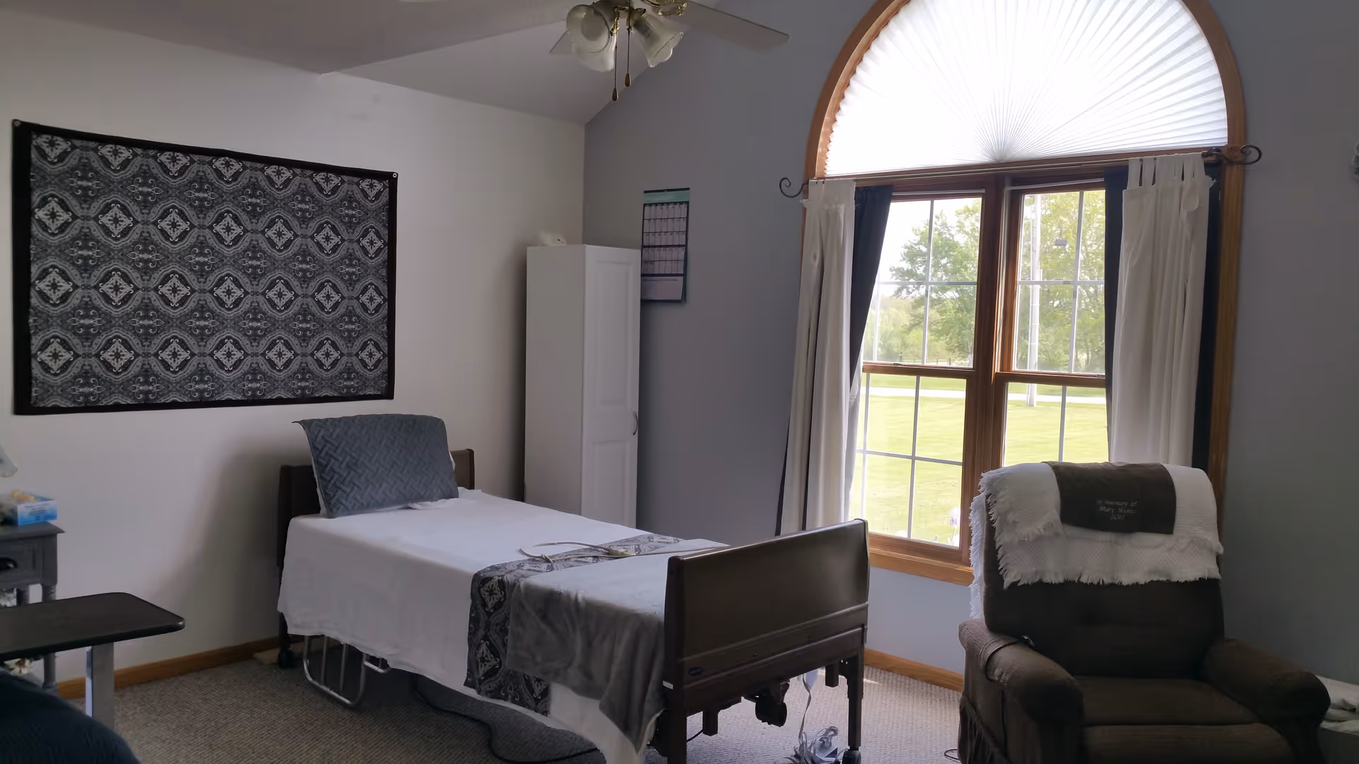 A bedroom in a senior facility with a bed, wardrobe, and arched window.