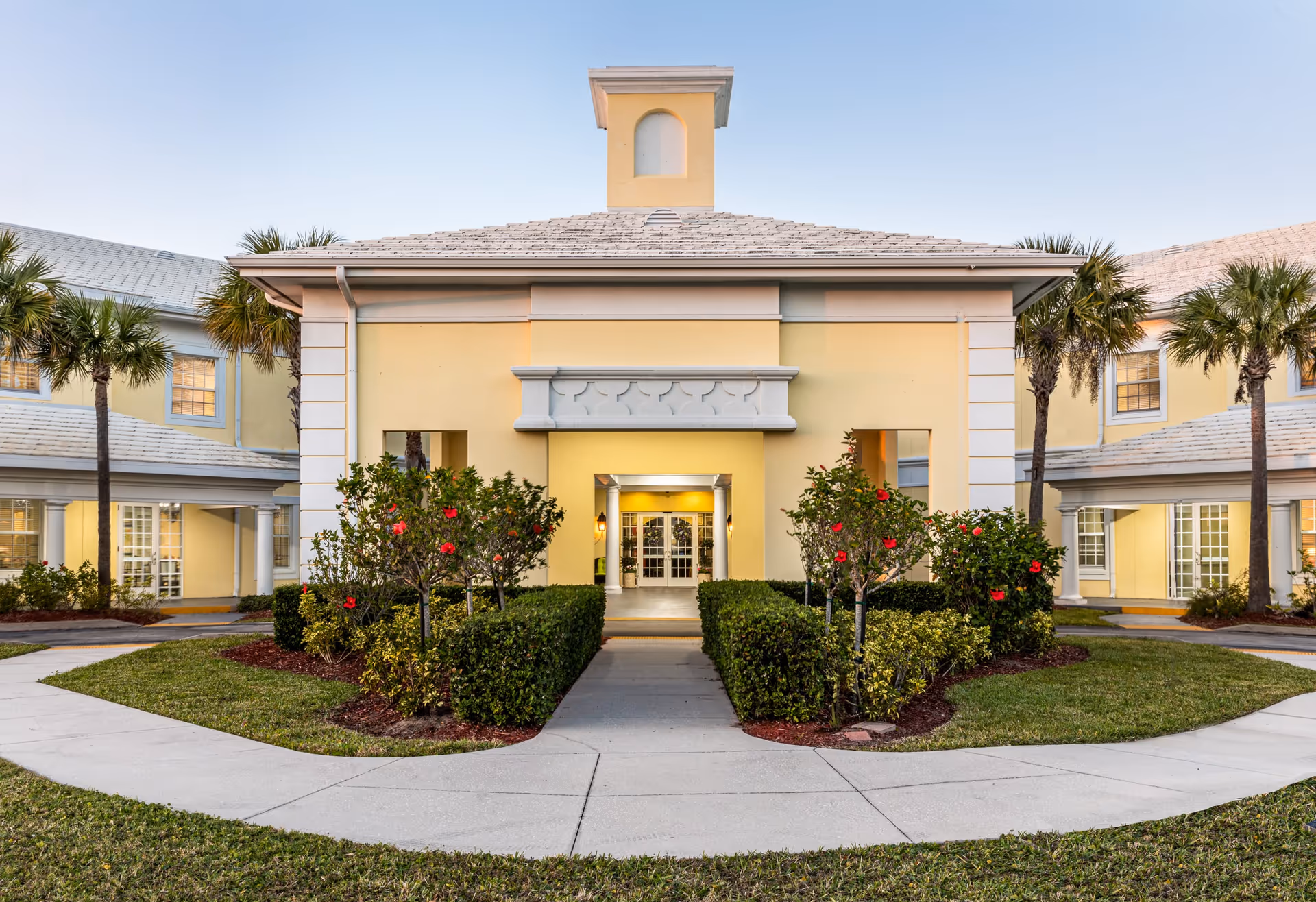 Front exterior view of a yellow assisted living facility building with a symmetrical design, palm trees, and neatly trimmed bushes with red flowers lining the walkway leading to the entrance.
