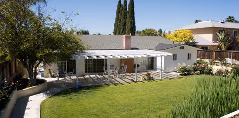 Single-story geriatric center with a shaded outdoor patio, green lawn, and surrounding landscaping.