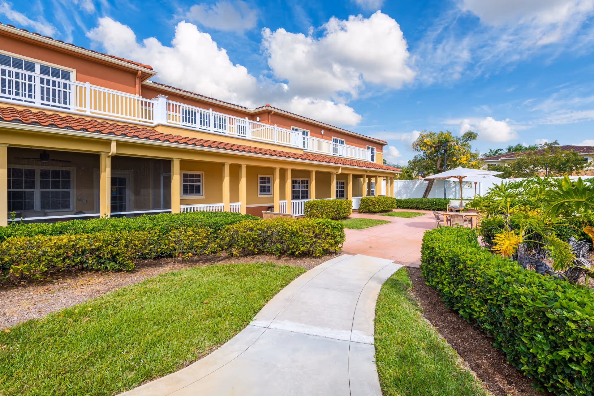 Outdoor view of Sonata Boynton Beach facility showing a two-story building with a red tile roof and yellow exterior walls. There is a curved concrete pathway leading through a well-maintained garden with green bushes and grass. Patio area with tables and umbrellas is visible on the right side under a partly cloudy blue sky.