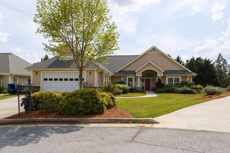 Front exterior of a single-story senior living building with a garage, entryway, tree and landscaped lawn.