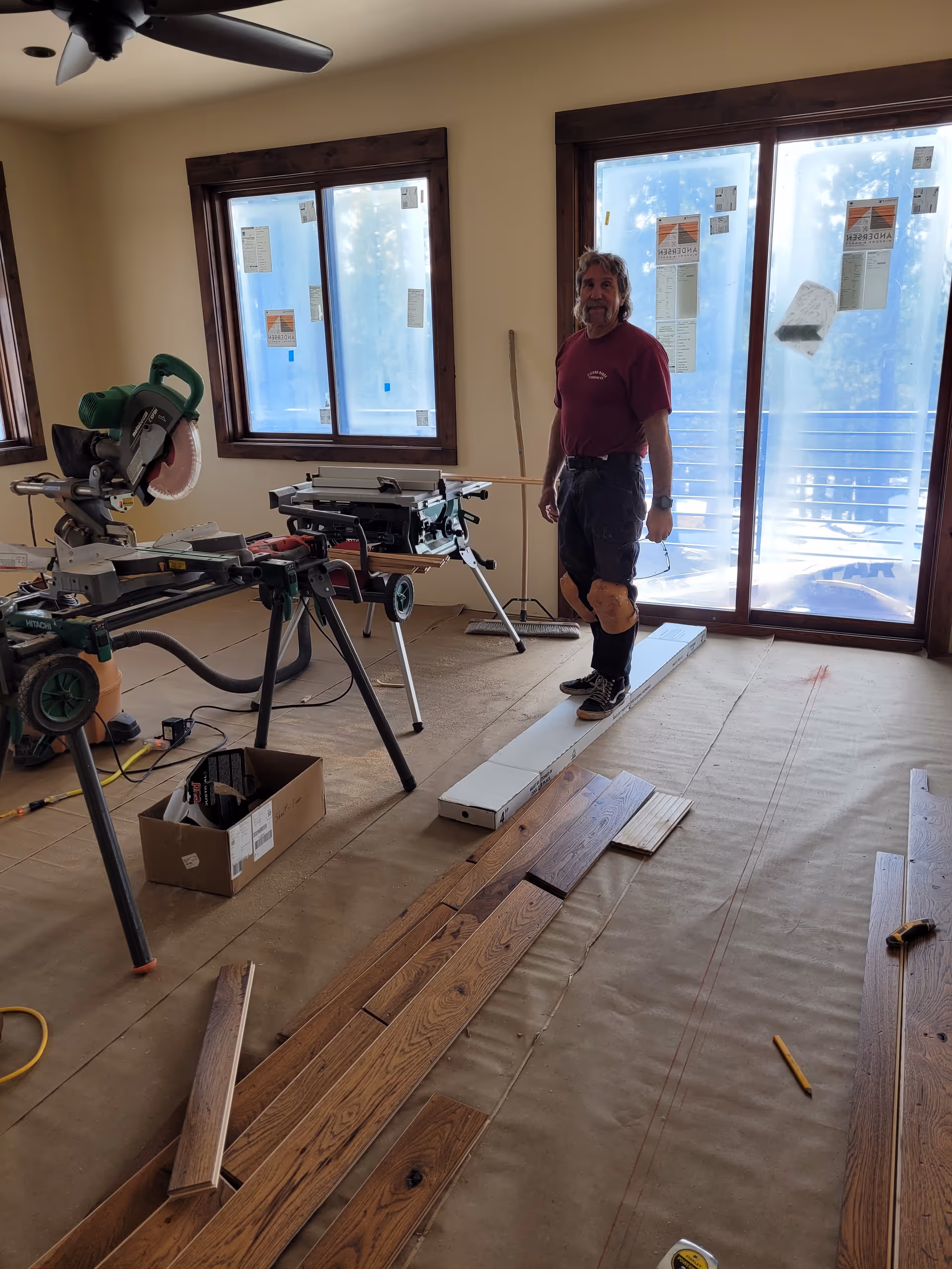 A man stands in a renovation interior with tools, planks, and a knee brace.