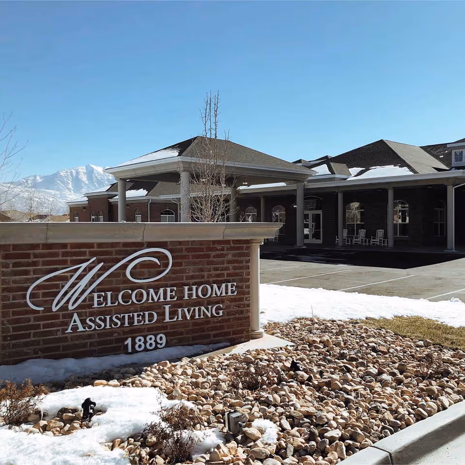 Brick sign reading 'Welcome Home Assisted Living 1889' in front of the facility entrance with snow-covered landscaping and mountains in the background.