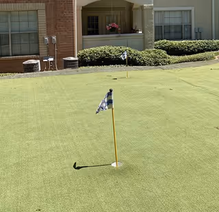 Putting green with two flagsticks in front of a residential building's ground-floor patios and shrubs.