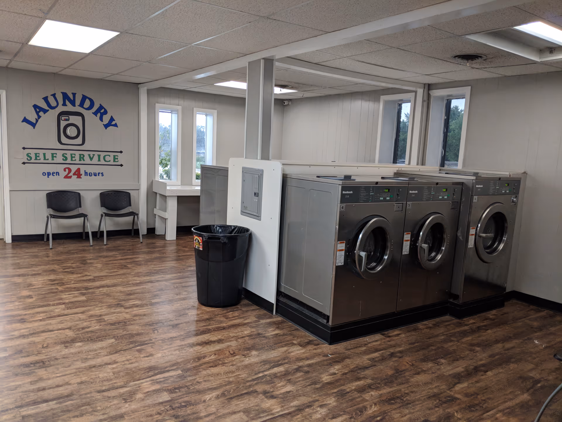 Laundry area with front-loading machines, a trash can, chairs, and a self-service sign open 24 hours.