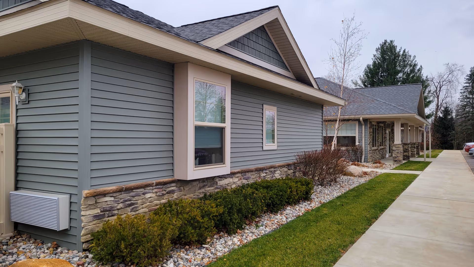 Exterior view of a single-story building with gray siding and stone accents, a covered porch supported by stone pillars, a concrete walkway, and landscaping with bushes and rocks along the side.