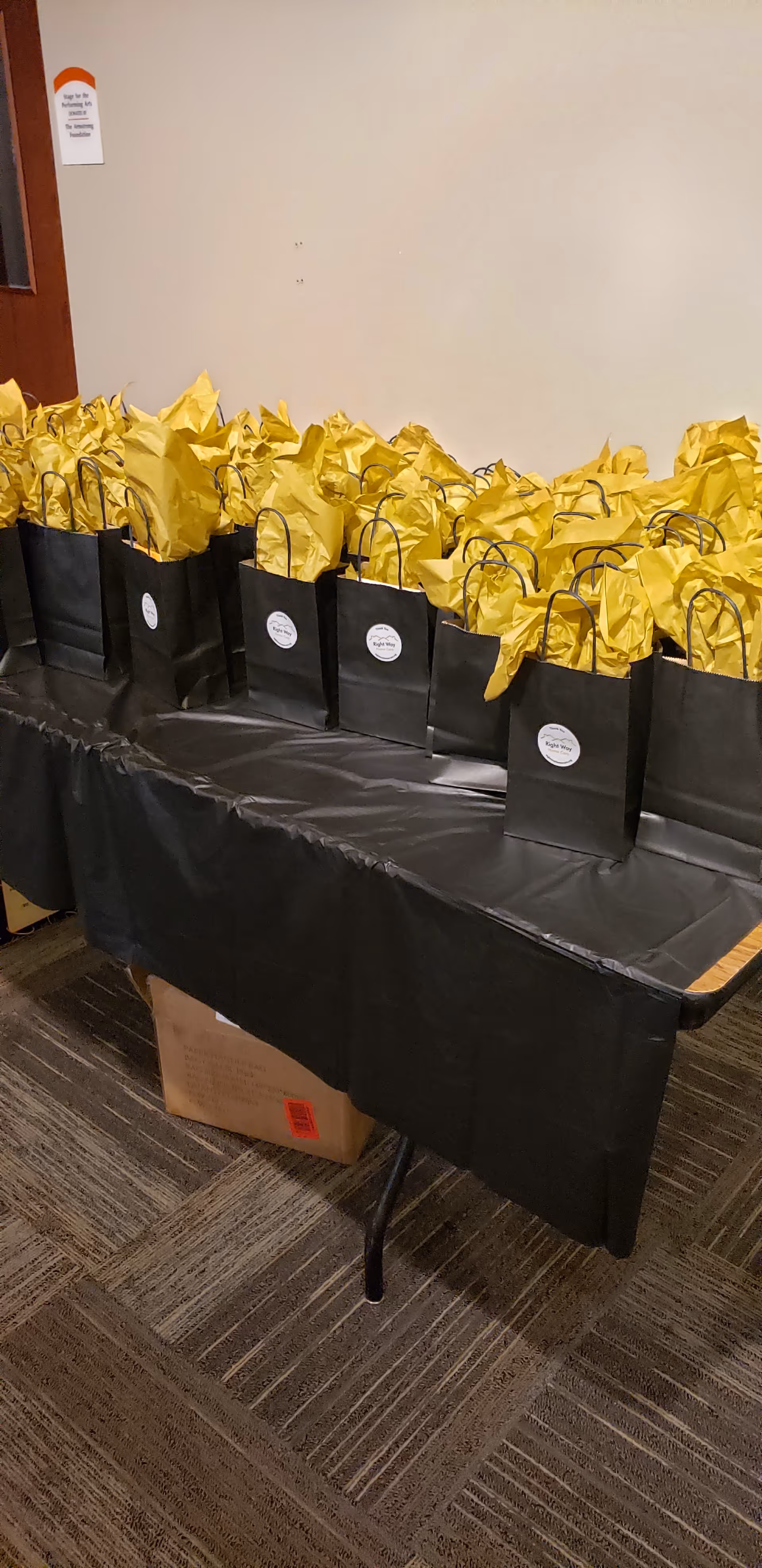 Row of black gift bags with yellow tissue on a long table in a hallway.