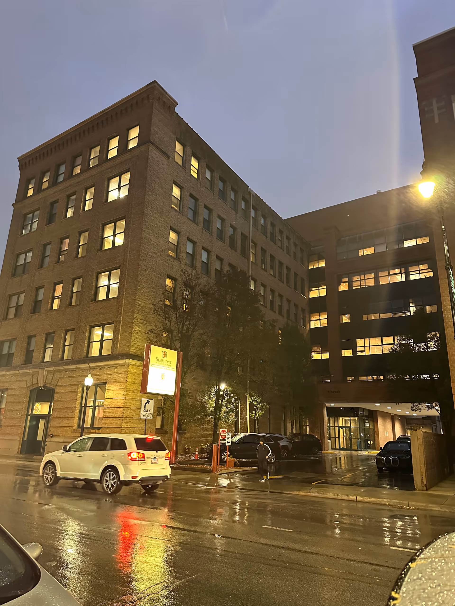 Exterior street-level view of a multi-story brick apartment building at dusk with many lit windows, parked cars and wet pavement.