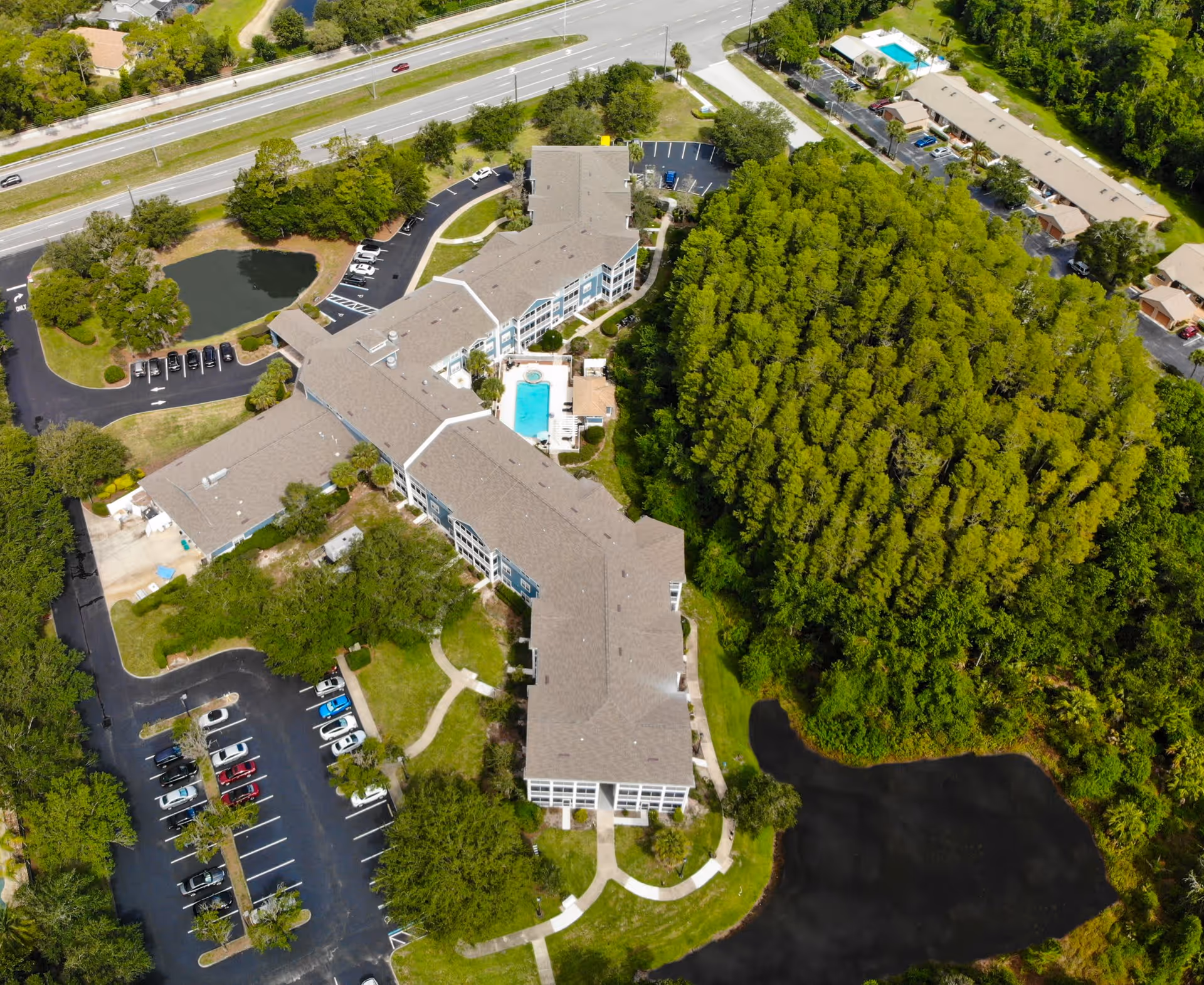 Aerial view of Addington Place of East Lake senior living facility showing a large building with a swimming pool in the center, surrounded by parking lots, trees, and two small ponds. A highway is visible in the background.