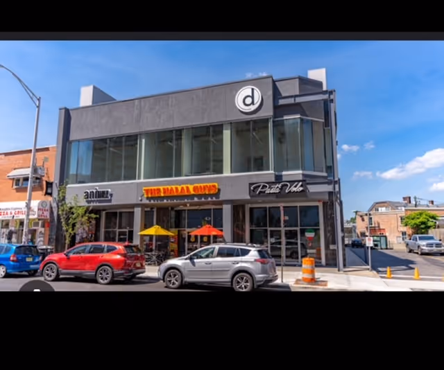 Two-story modern storefront building with large glass windows, outdoor seating under umbrellas, and parked cars.