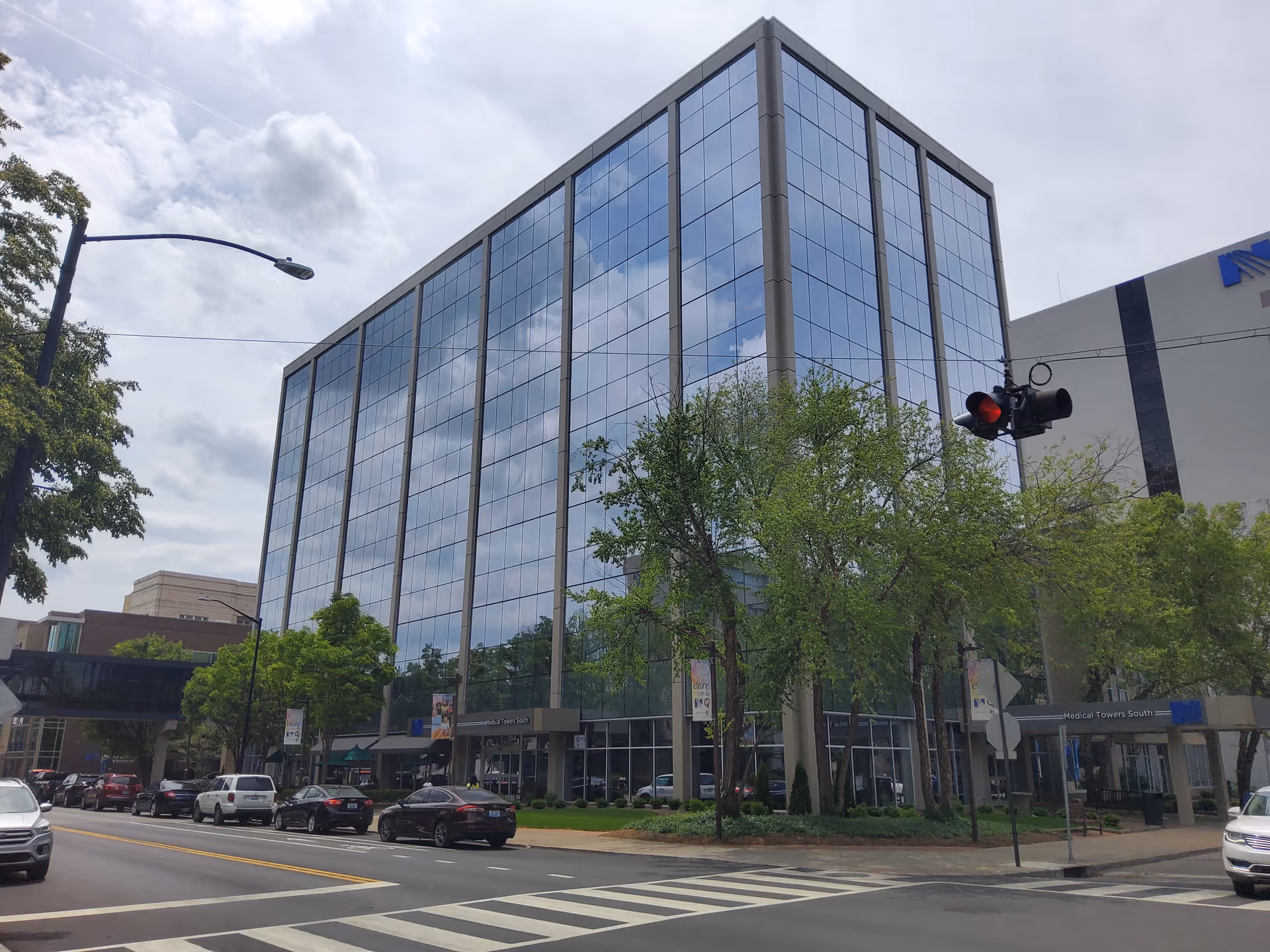 Glass-fronted, multi-story building with trees, cars, and a crosswalk along a city street.