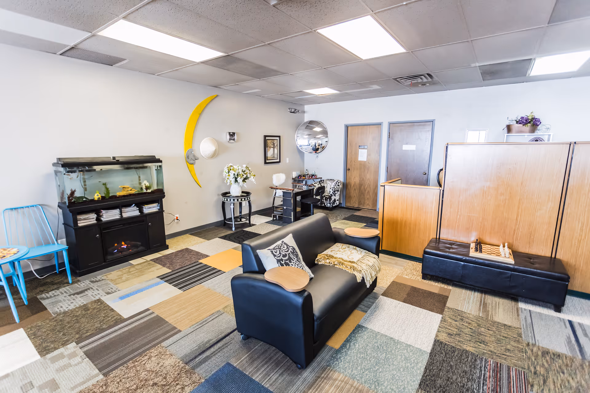 Cozy senior living lounge with a black sofa, fish tank, chessboard, and multicolor carpet.