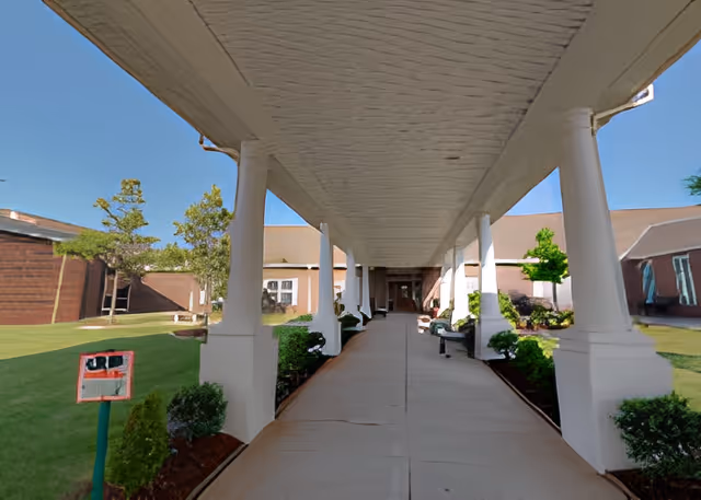 Covered entrance walkway with white columns leading to a building entrance, flanked by benches and landscaped lawns.