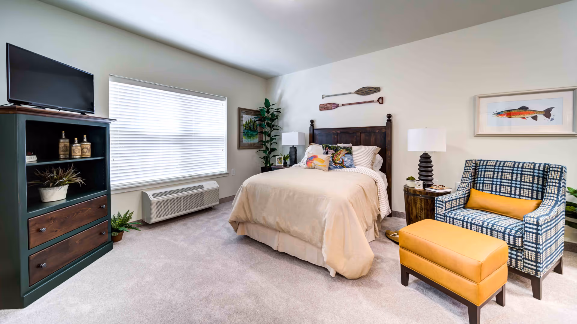 Bright furnished bedroom with a bed, patterned armchair and ottoman, TV cabinet, and window with blinds.